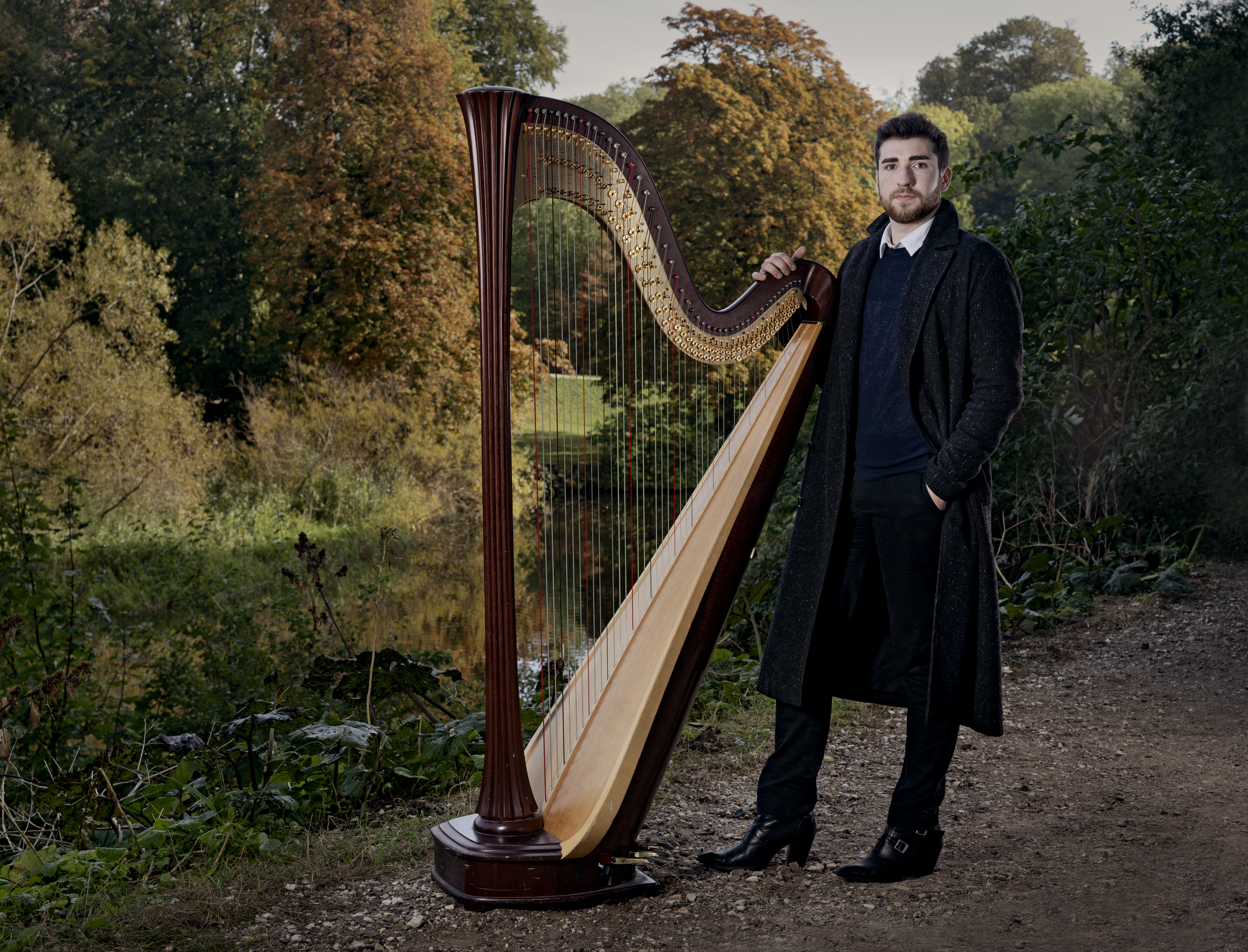 Harpenist Zachary Hatcher iført efterårsfrakke i skoven står med sin store og betagende harpe i grønne omgivelse.