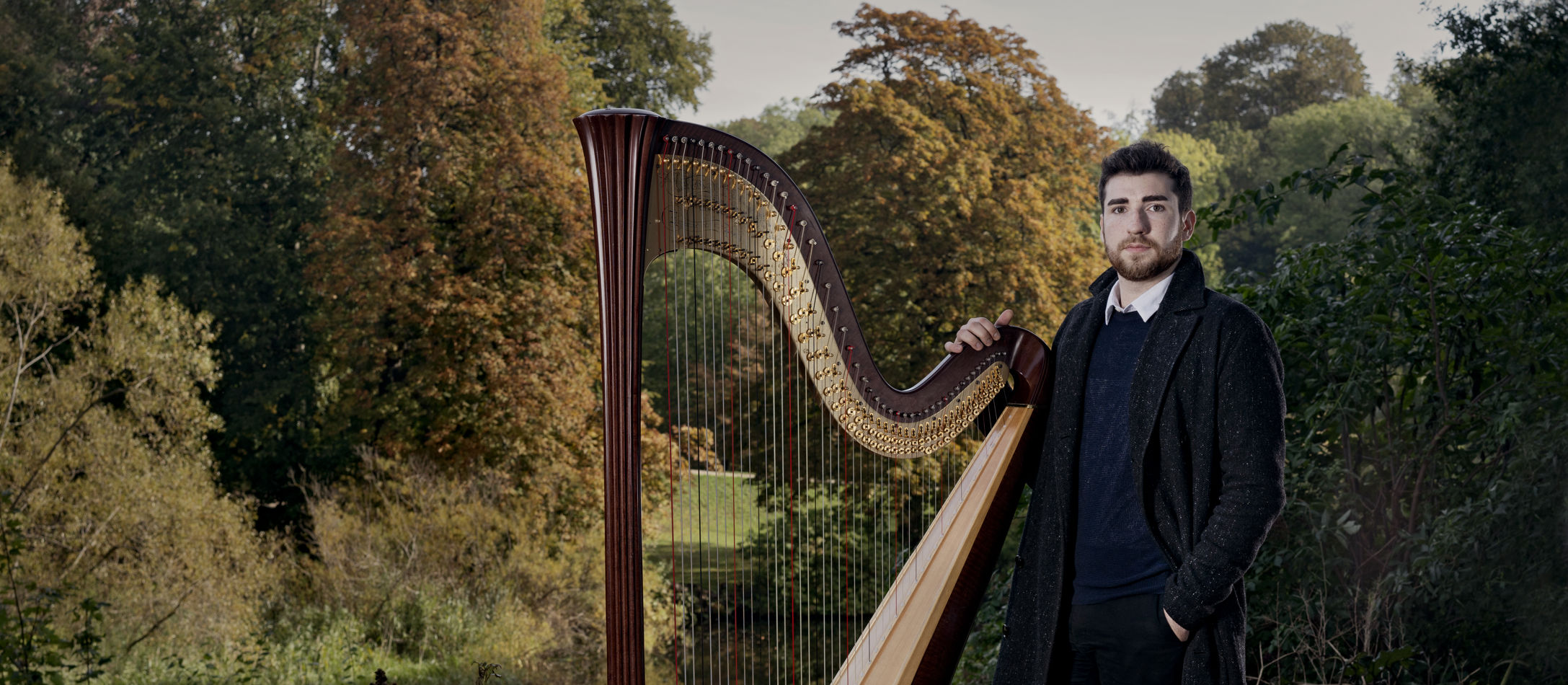 Harpenist Zachary Hatcher iført efterårsfrakke i skoven står med sin store og betagende harpe i grønne omgivelse.
