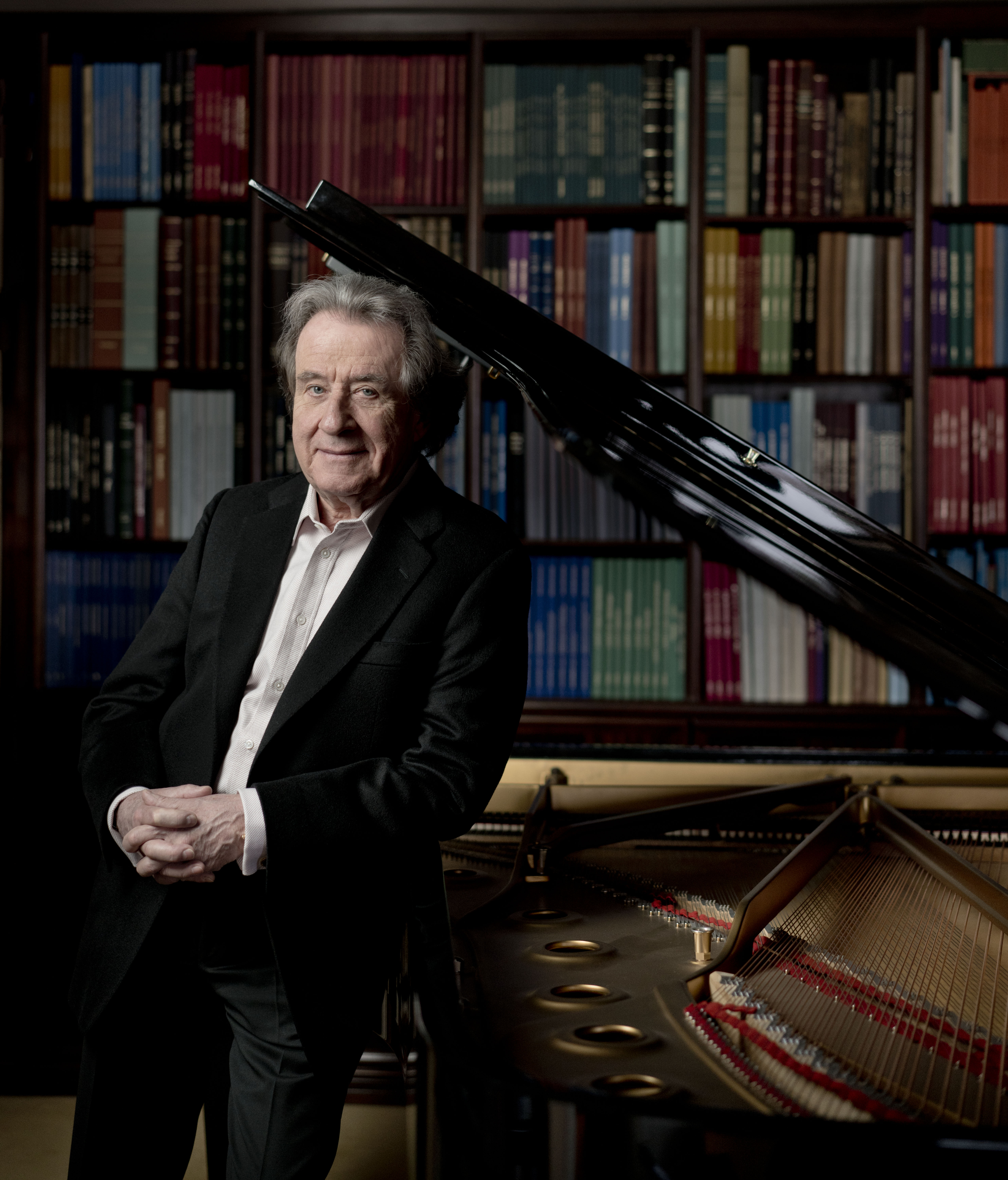 Rudolf Buchbinder læner op ad reflekterende klaver foran stor boghylde iført klassisk jakkesæt.