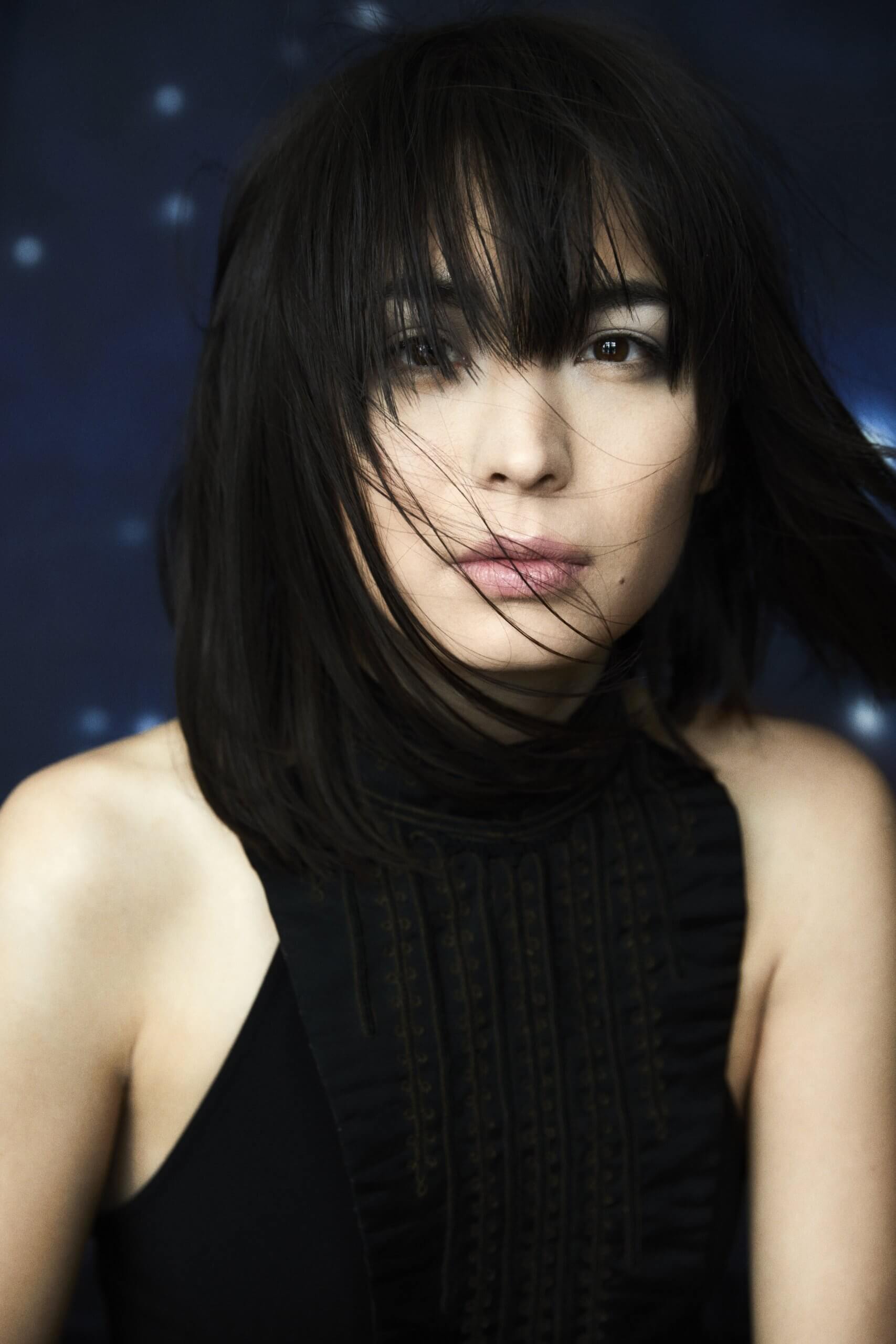 Alice Sara Ott med mørkt pandehår, der falder foran dybe, brune øjne med bare skuldre og høj hals på bluse med blå baggrund 