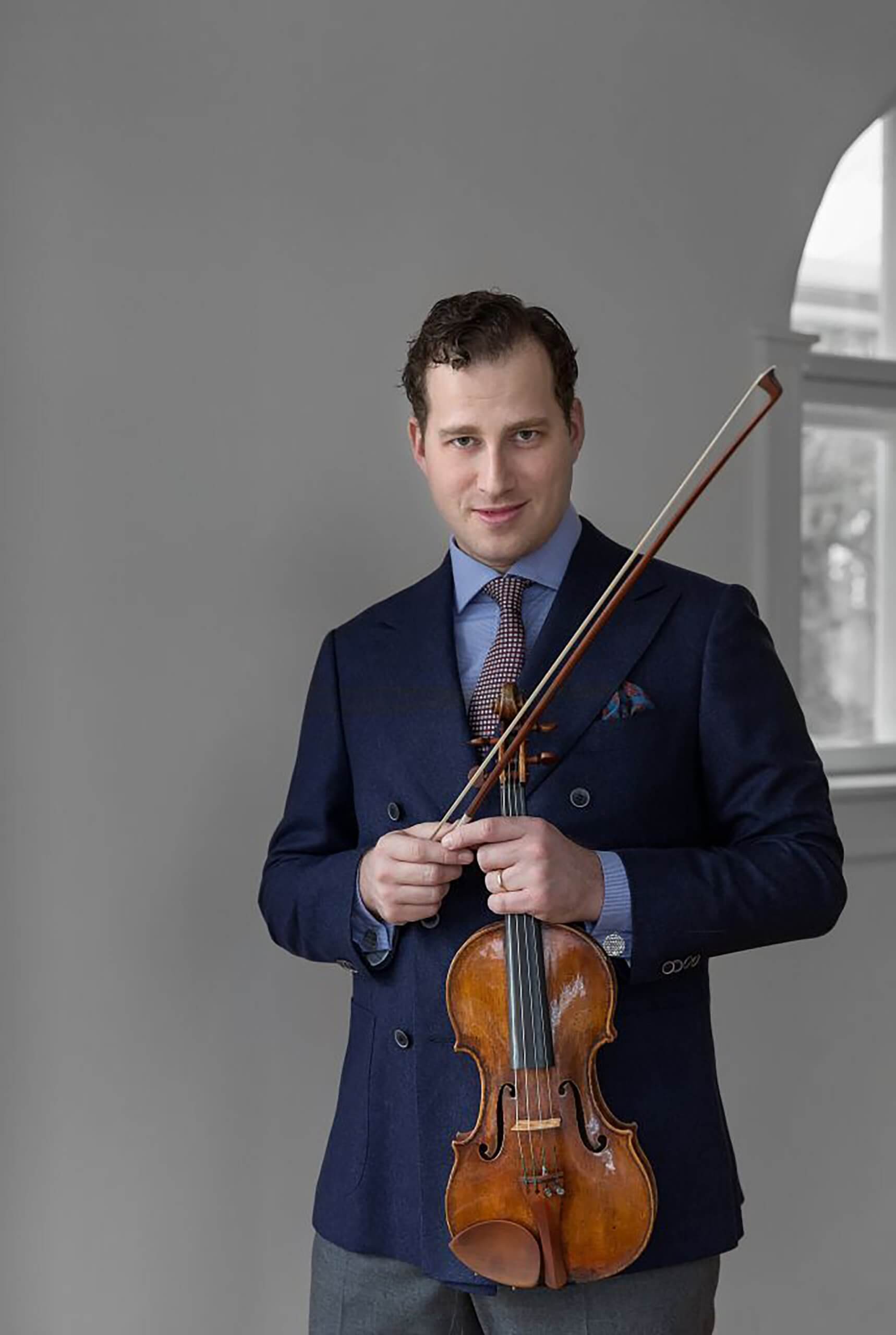 Nikolaj Szeps Znaider med fin violin i hånden foran hvid væg og vindue iført lyseblå skjorte og mørkeblå jakke udover og gå bukser. 