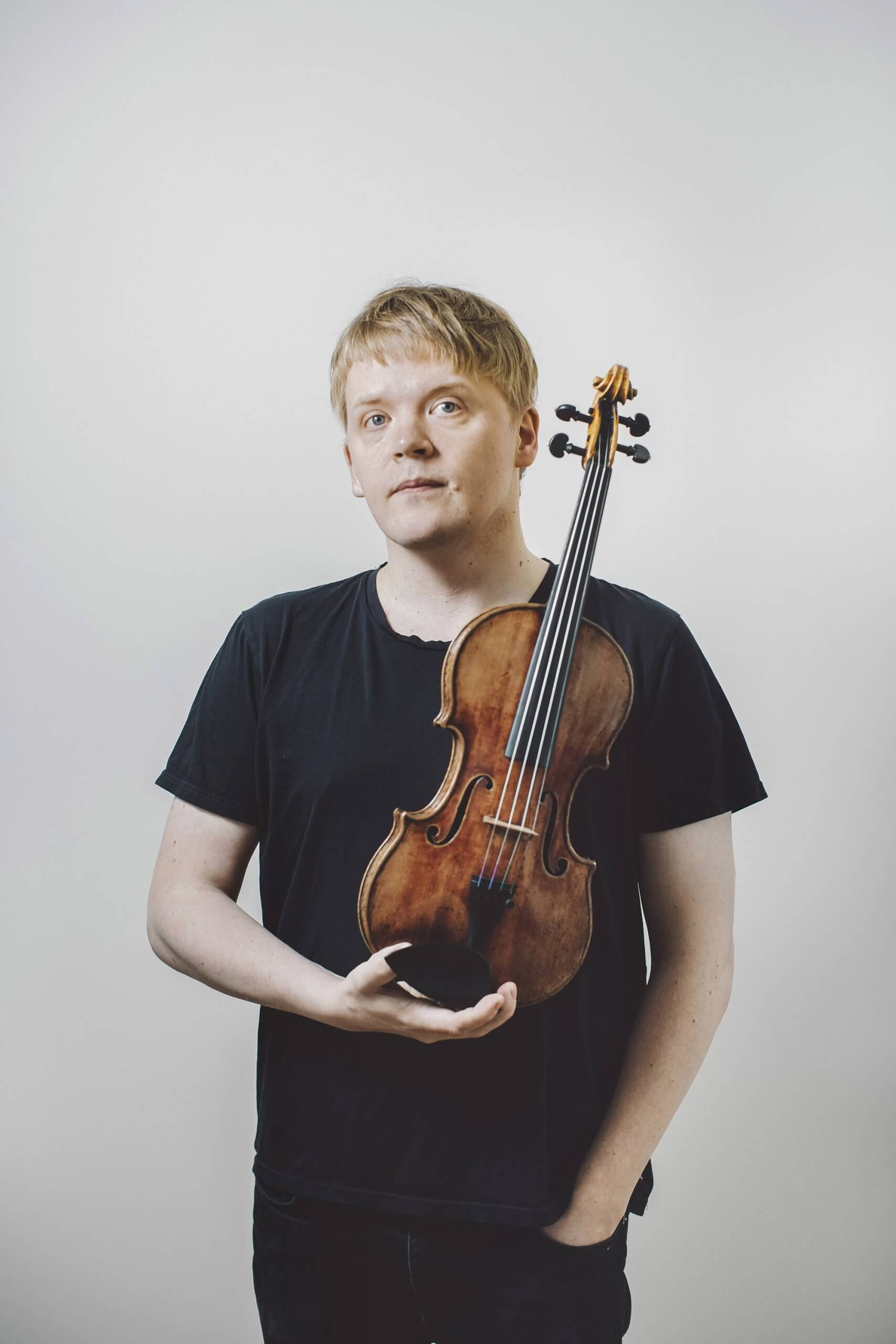 Pekka Kuusisto iført sort t-shirt og sorte bukser foran hvid væg holder sin fine violin i den ene hånd den anden i lommen. 