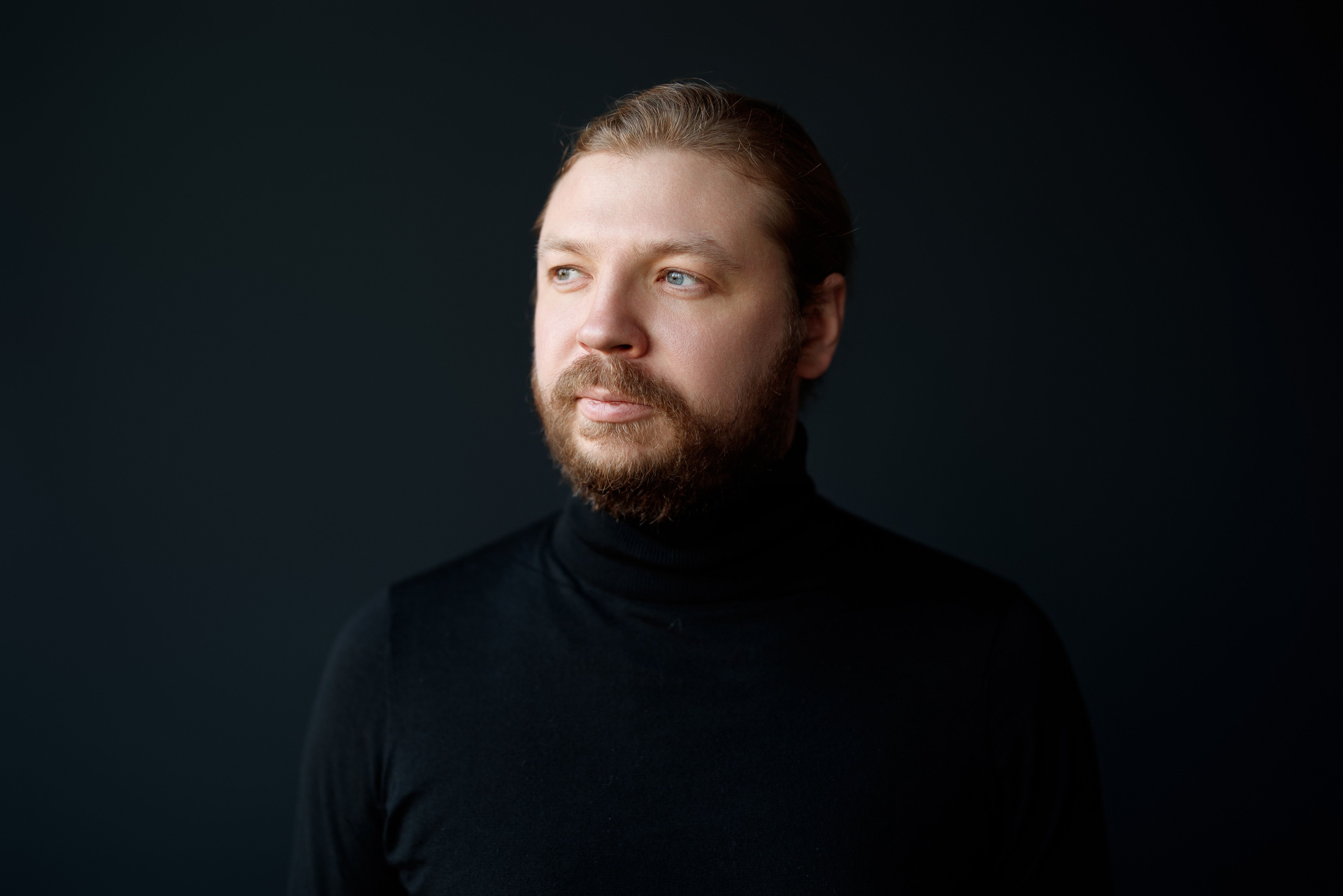 Pianisten Denis Kozhukhin med sort halskrave oh rødligt hår og skæg og blå øjne kigger mod sin højre foran sort baggrund. 