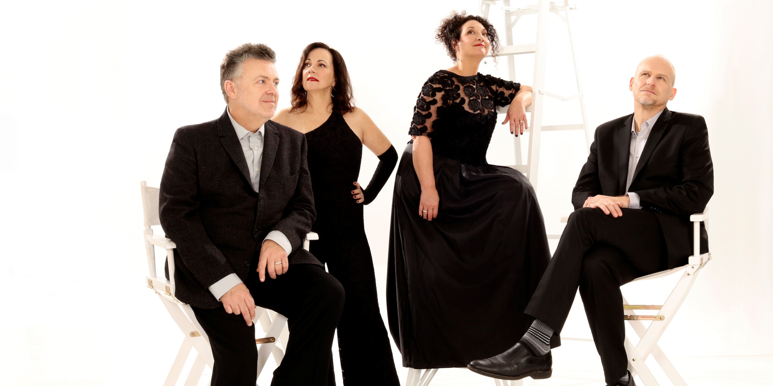 Fire sangere i pænt sort tøj, stå og sidder ved to hvide stole og en hvid stige.