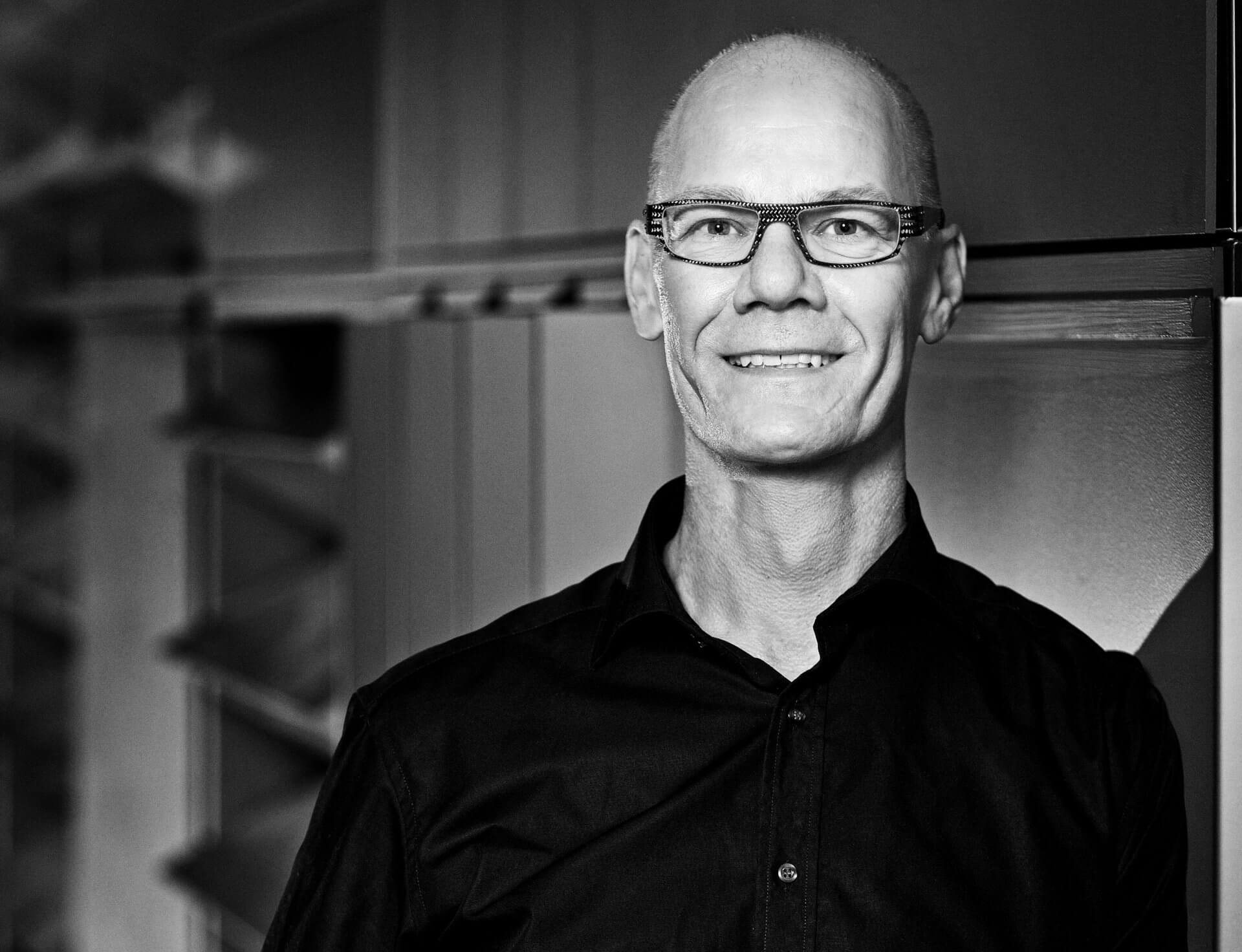 Poul Emborg med briller storsmiler iført sort skjorte. 