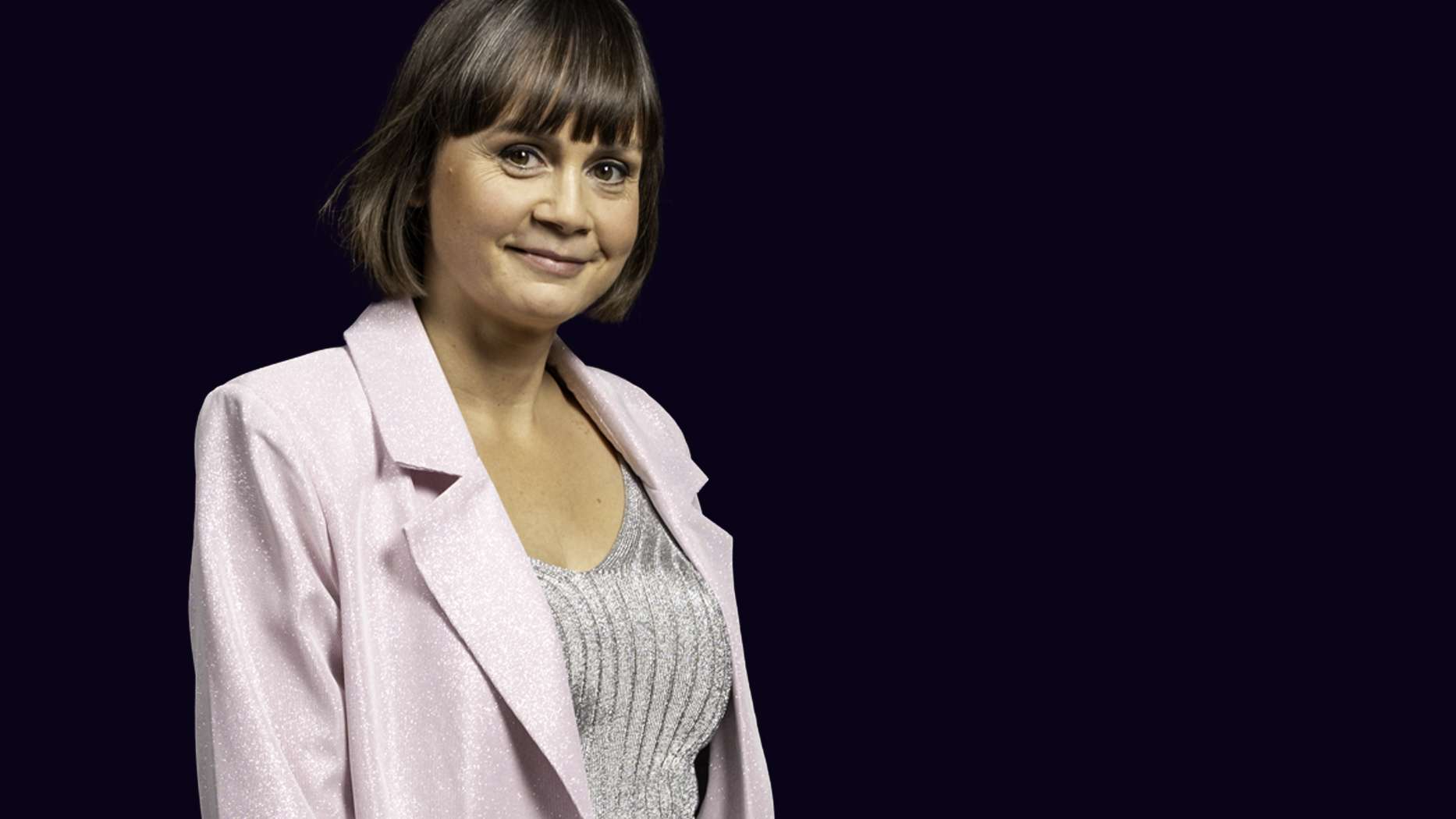 Smilende vært Maria Månson iført rosafarvet jakkesæt og glimmertop på sort baggrund