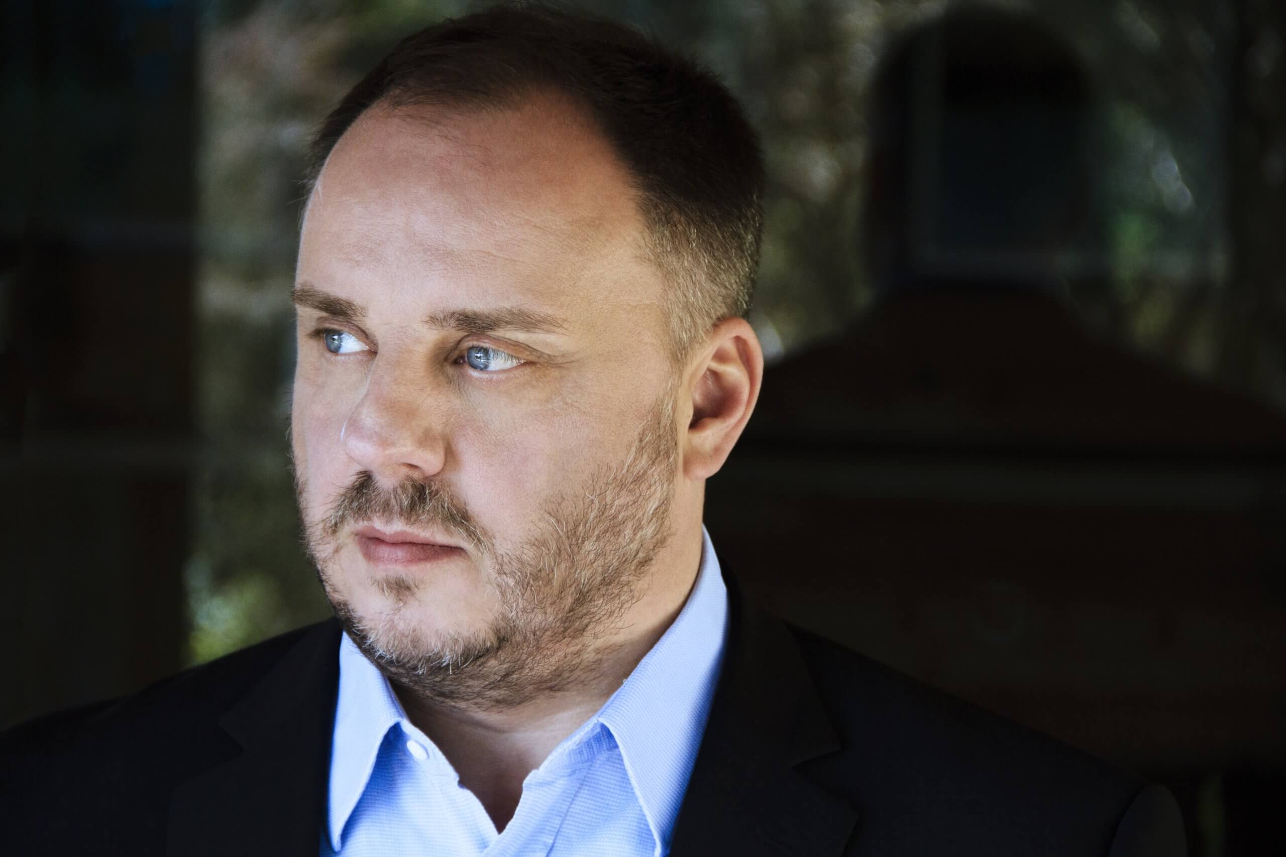 Matthias Goerne kigger med sine blå øjne mod sin højre iført lyseblå skjorte og sort jakke udover på sløret baggrund. 
