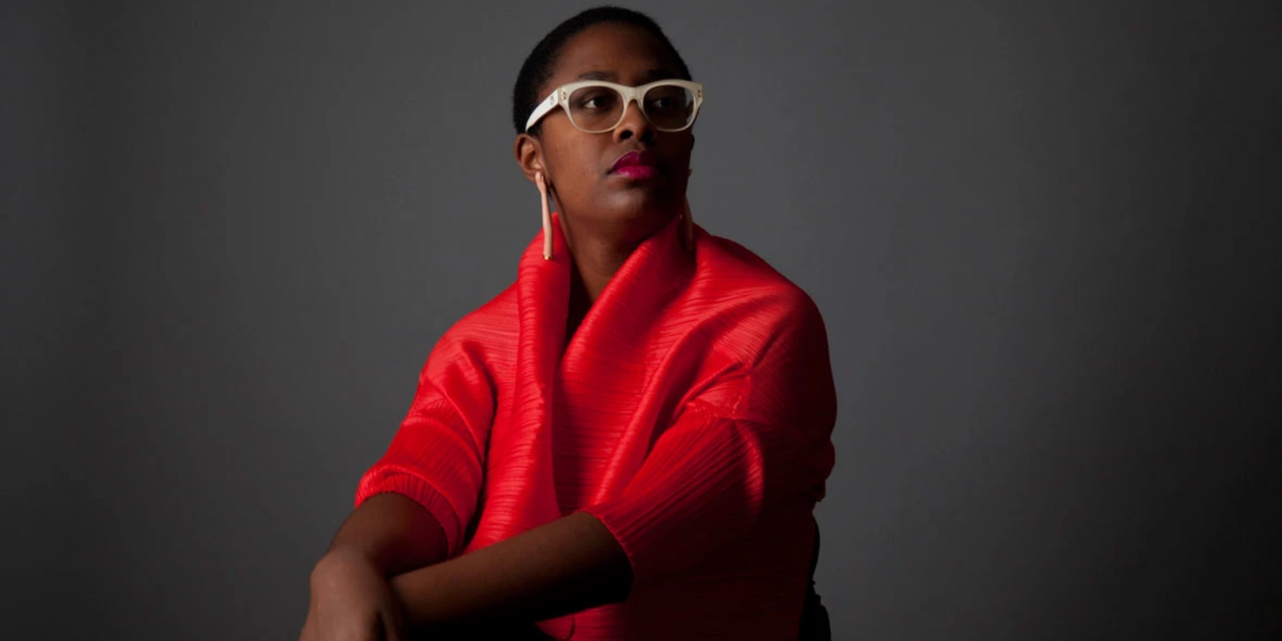 Kvinde i rødt med hvide briller, sidder foran grå baggrund.