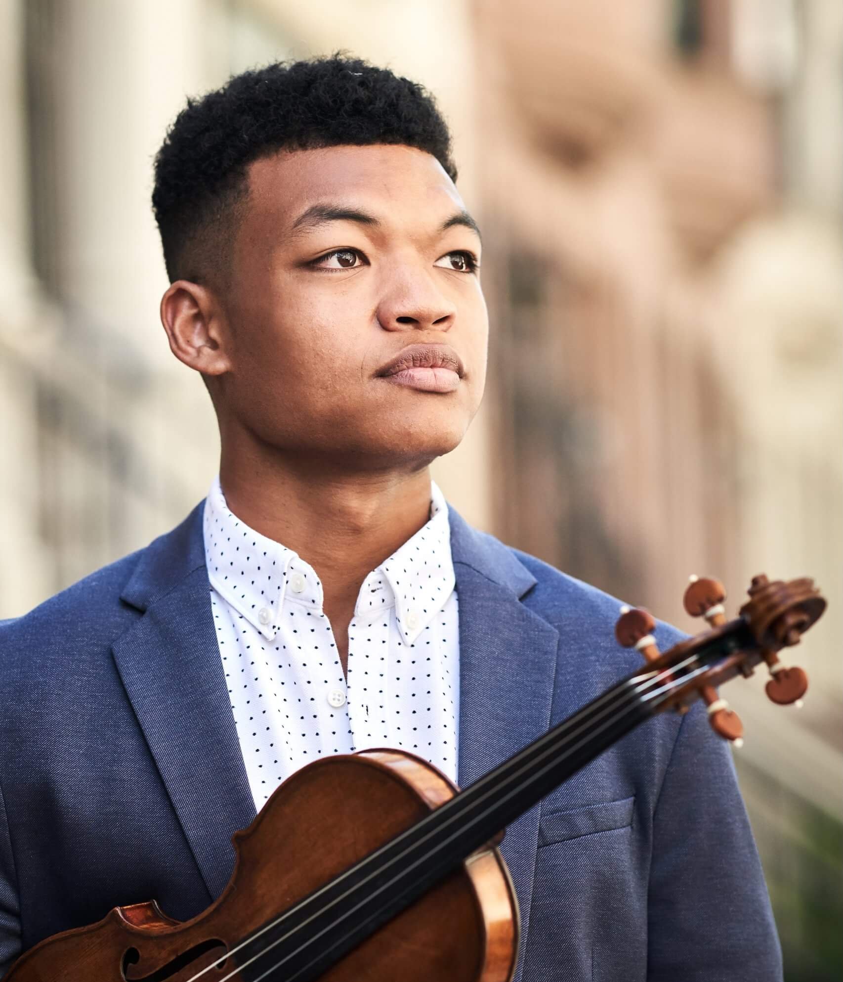Mørkhudede violinist Randall Goosby iført sortprikket hvid skjorte og grålig jakke foran sløret baggrund med sin viollin.
