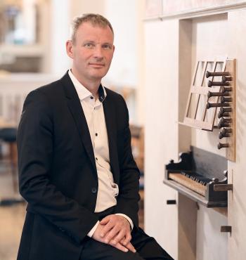 Organist Søren Christian Vestergaard siddende foran lille orgel iført hvid skjorte og sort jakke smiler let. 