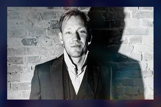 James Sherlock dirigent til Gaming X Symphony i sort/hvid foran murstensmur. 