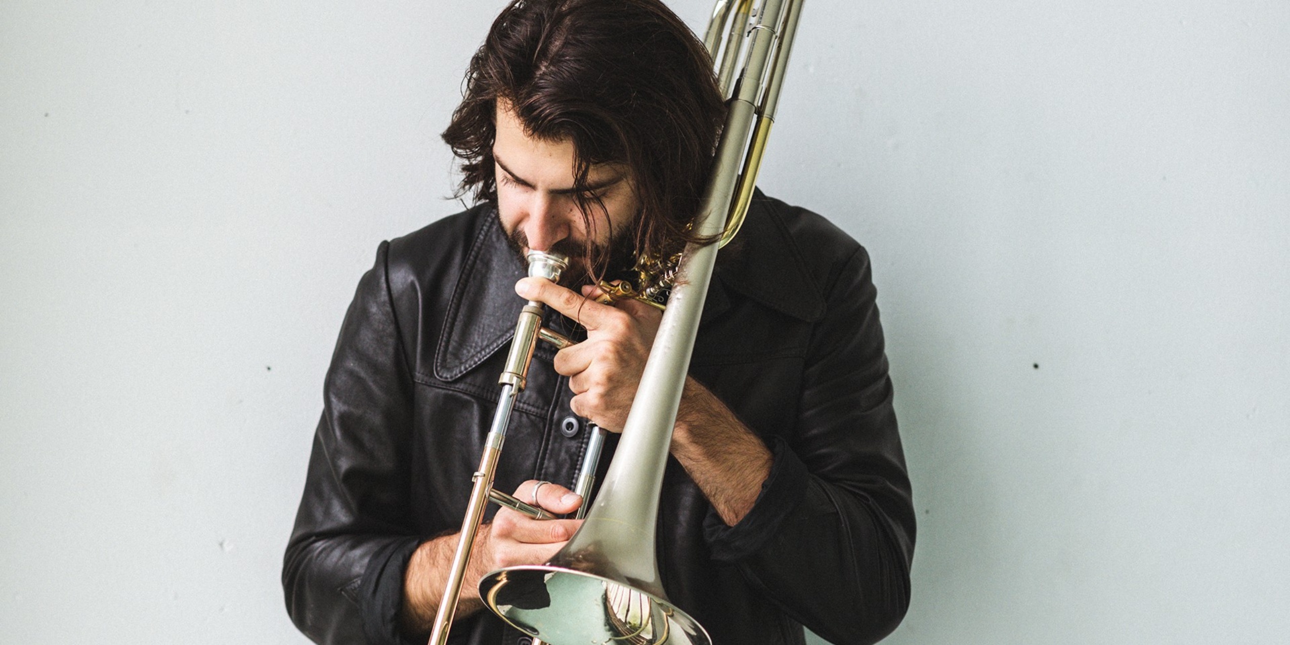 Mand der spiller basun i sort læderjakke foran hvid baggrund.