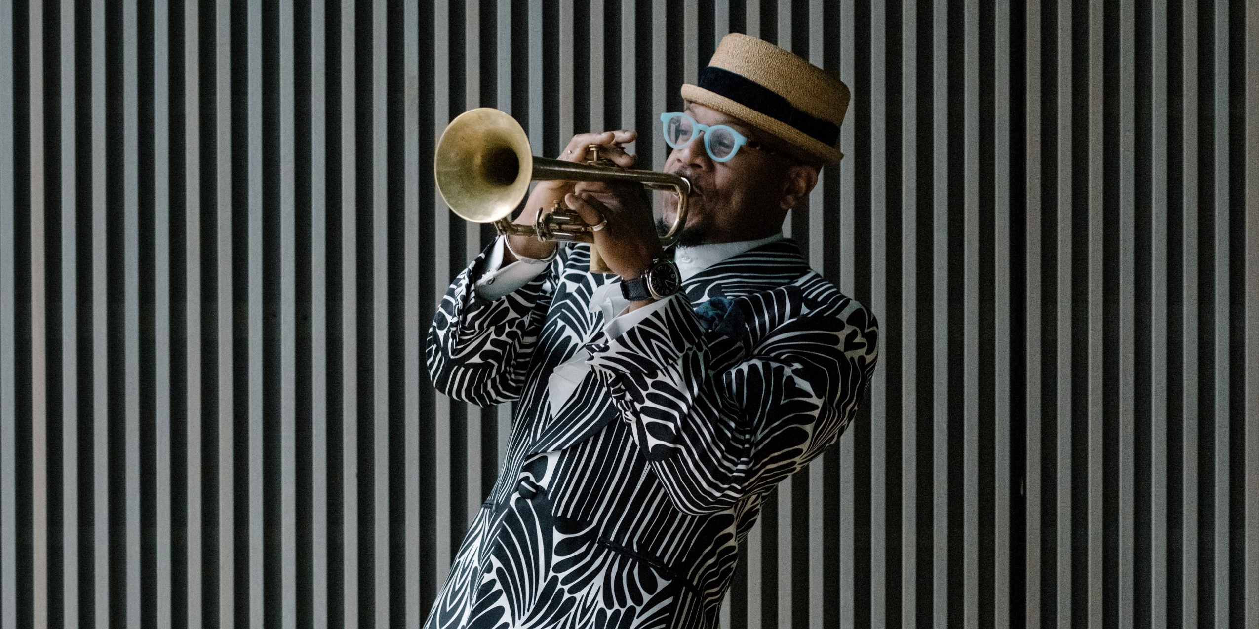 Mand i sort og hvid geometrisk blazer og hat, spiller trompet foran en sort og hvid stribet væg.