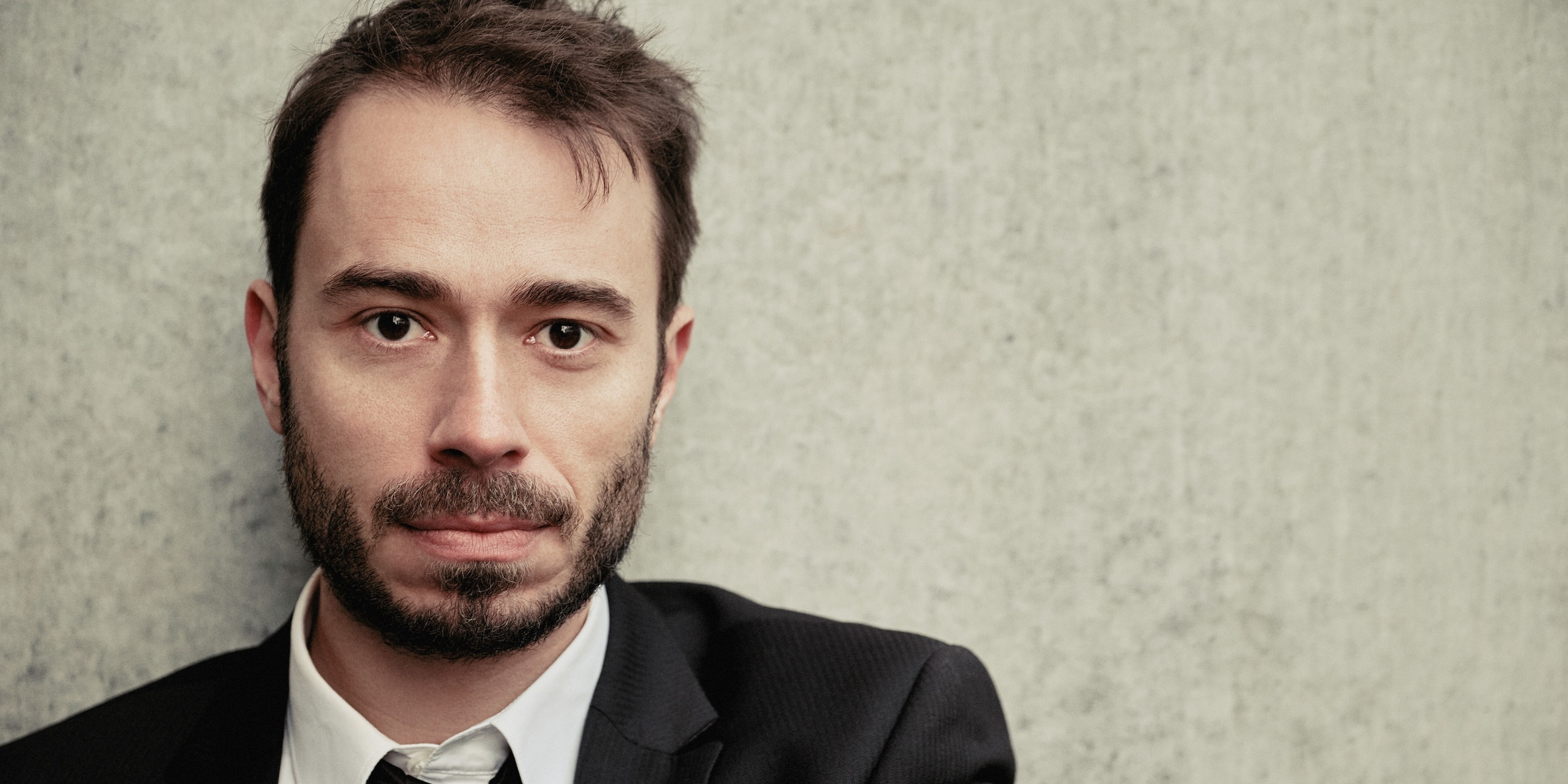 Mand sort blazer og hvid skjorte, kigger ind i kameraet foran en grå betonvæg.