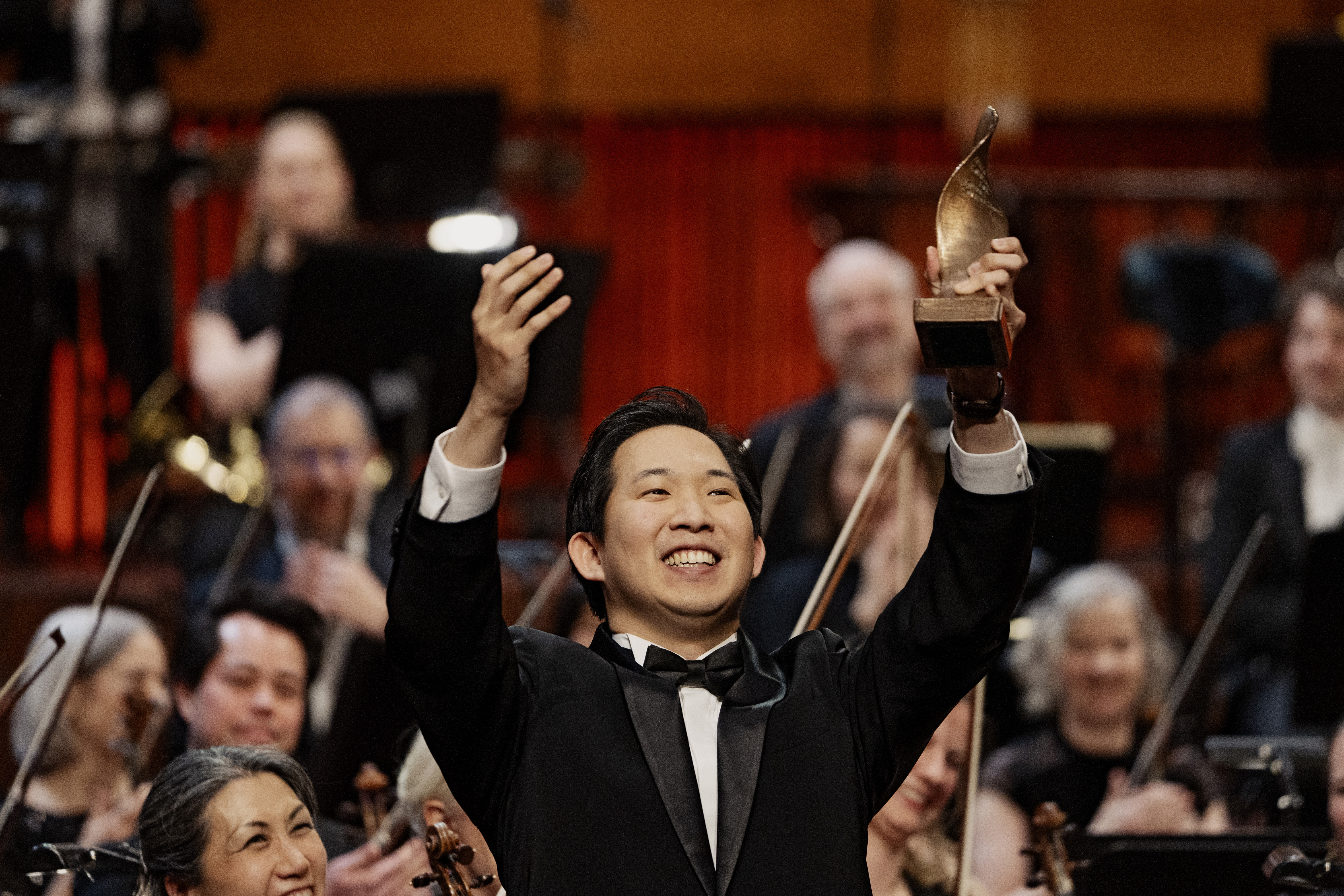 Storsmilende sydkoreanske Samuel S. Lee i klassisk suit med statuette i den ene hånd og stort orkester i baggrunden