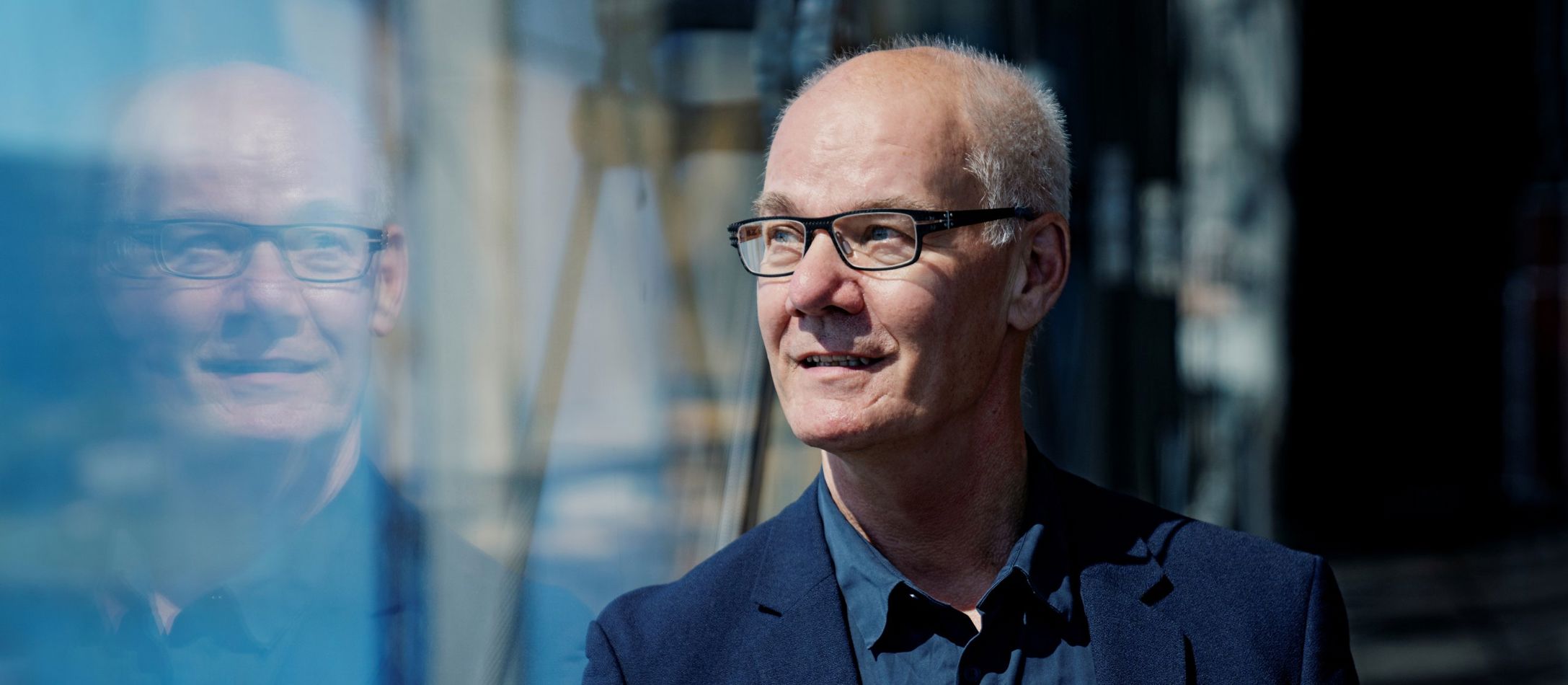 Poul Emborg med briller læner sig op ad glasrude iført blålig afslappet jakke og skjorte og reflekteres i spejlet.
