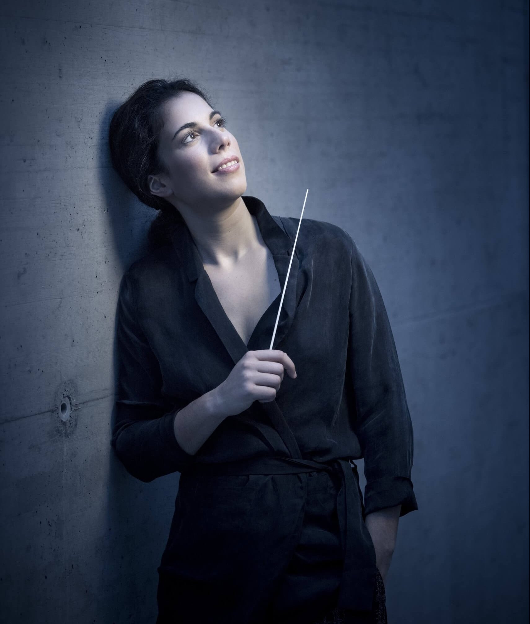Marie Jacquot med dirigentstang i den ene hånd smiler mens hun læner sig op ad grå mur og kigger opad. 