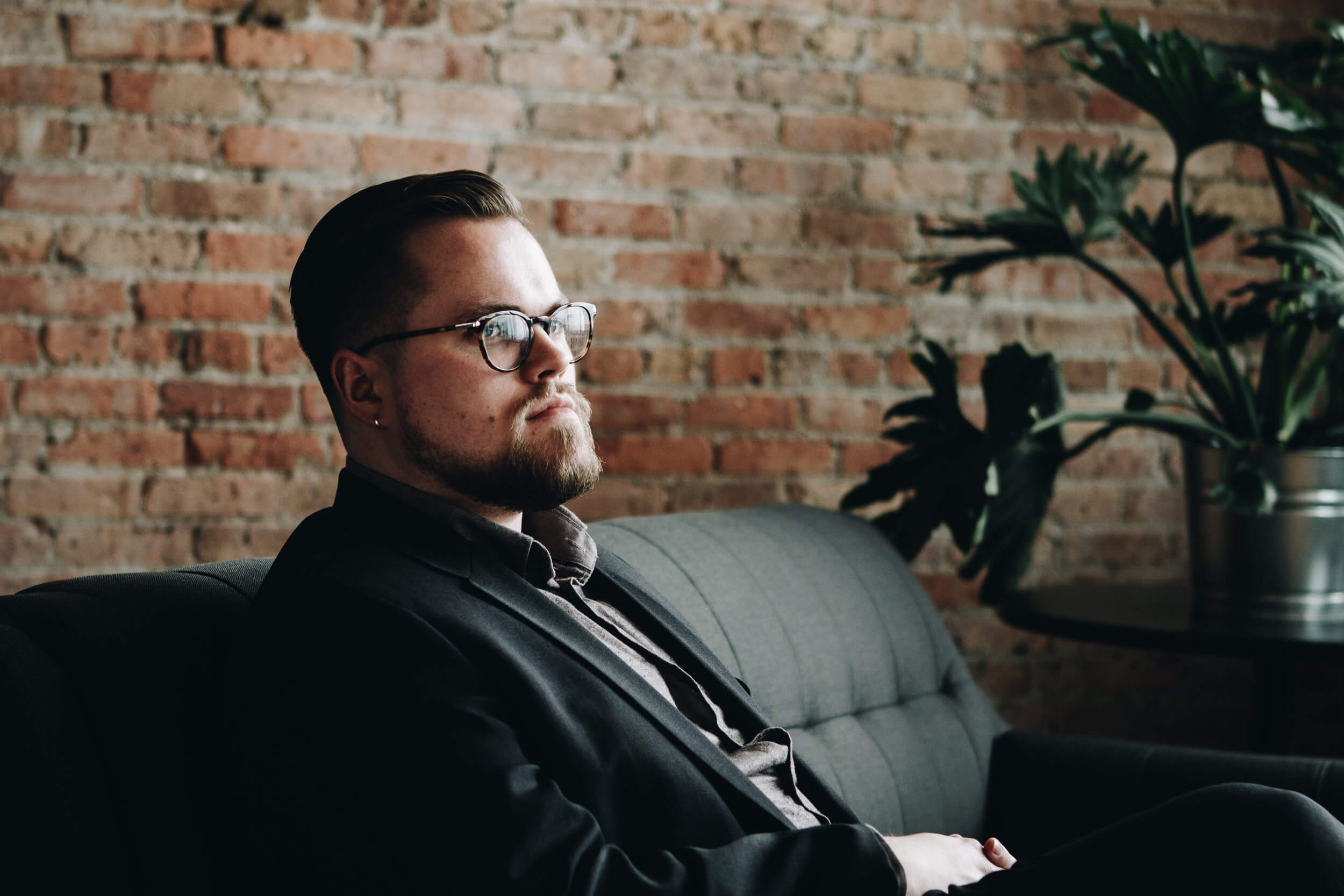 James Ley sidder i sort sofa iført briller med flot skæg foran rød murstensvæg indendørs 