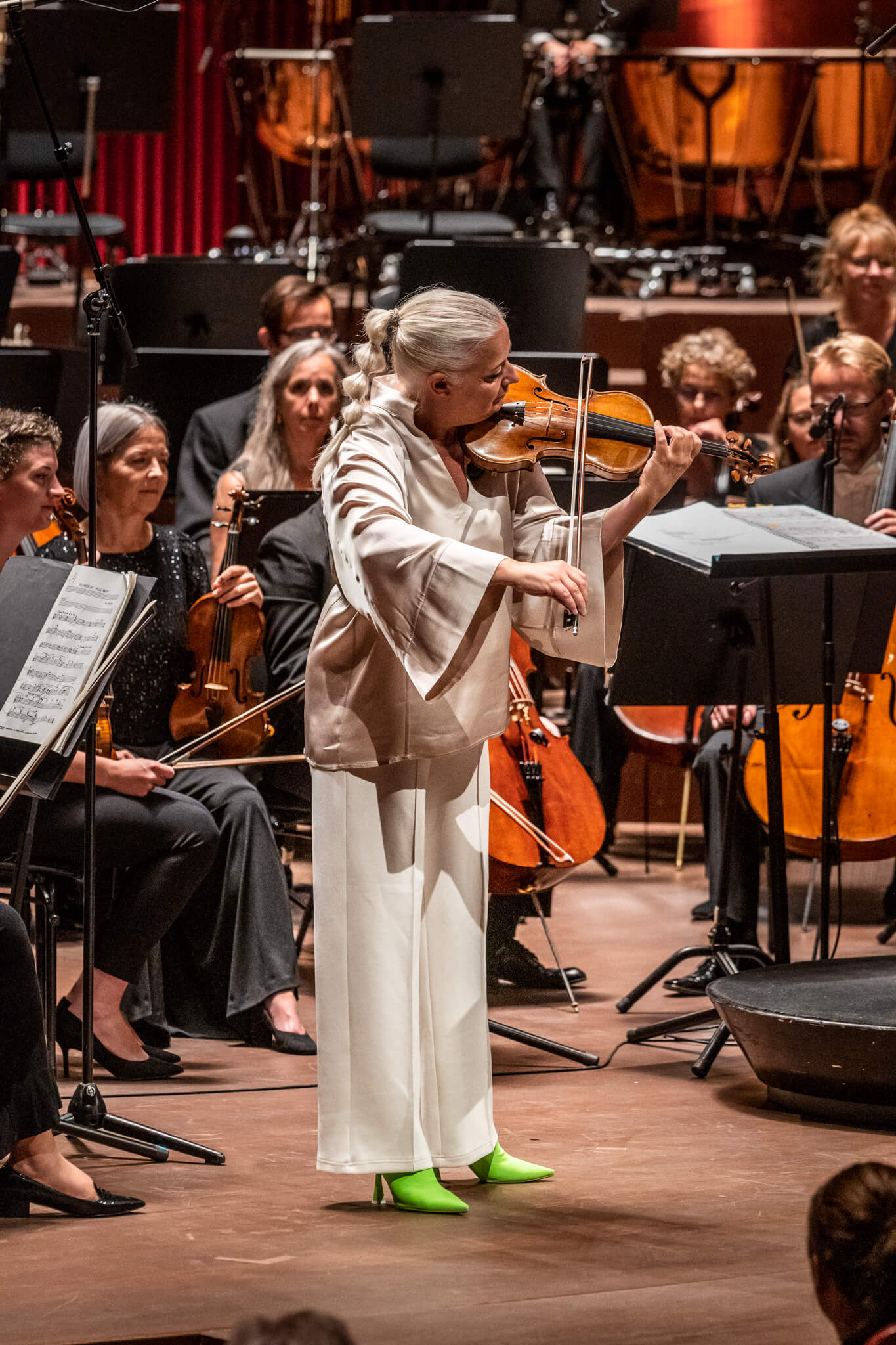 Christina Åstrand står og spiller violin i hvidt sæt tøj foran stort orkester, der sidder ned og venter.