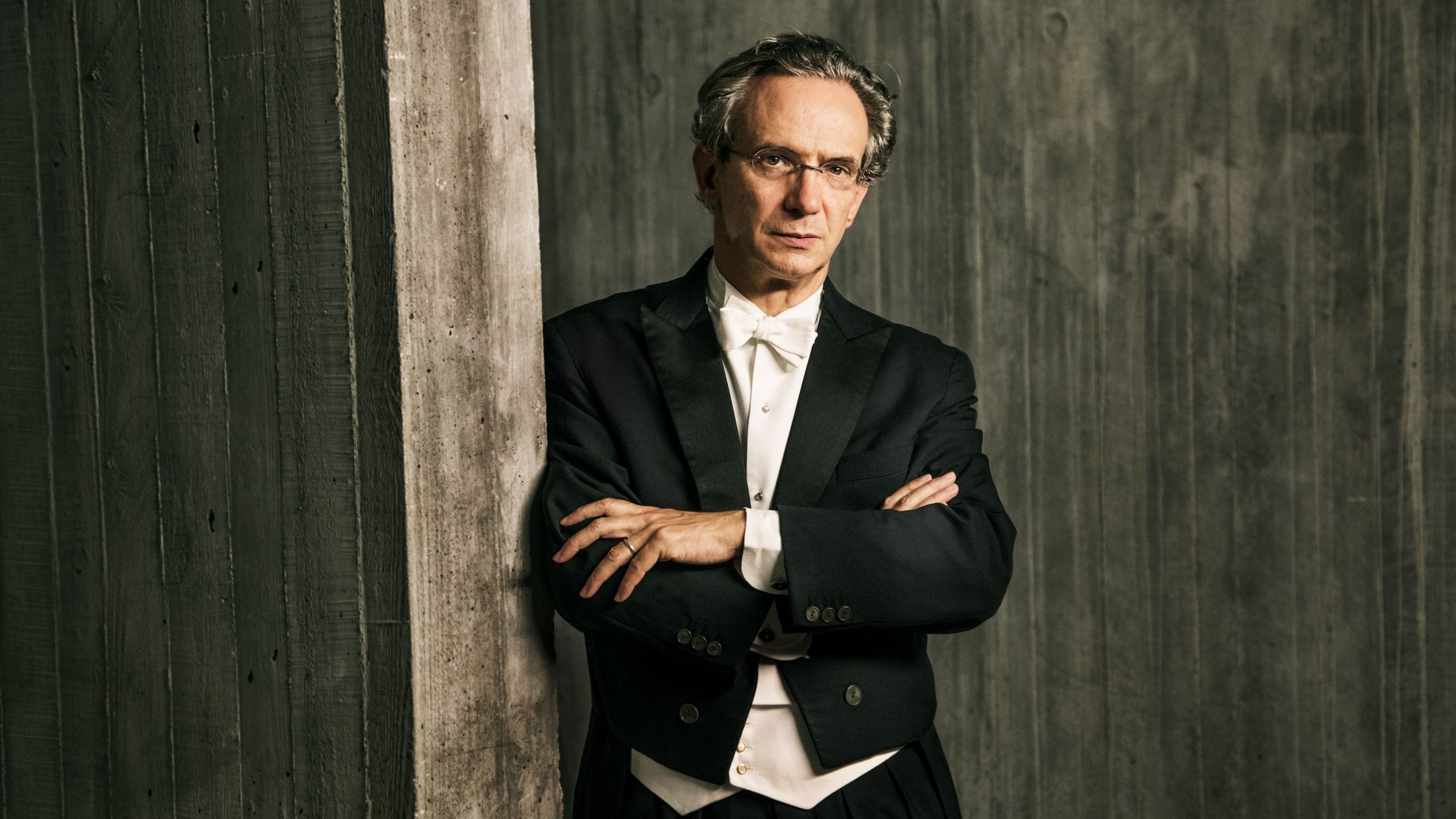 Fabio Luisi med kortlagte arme iført elegant gammeldags habit læner sig opad træværk