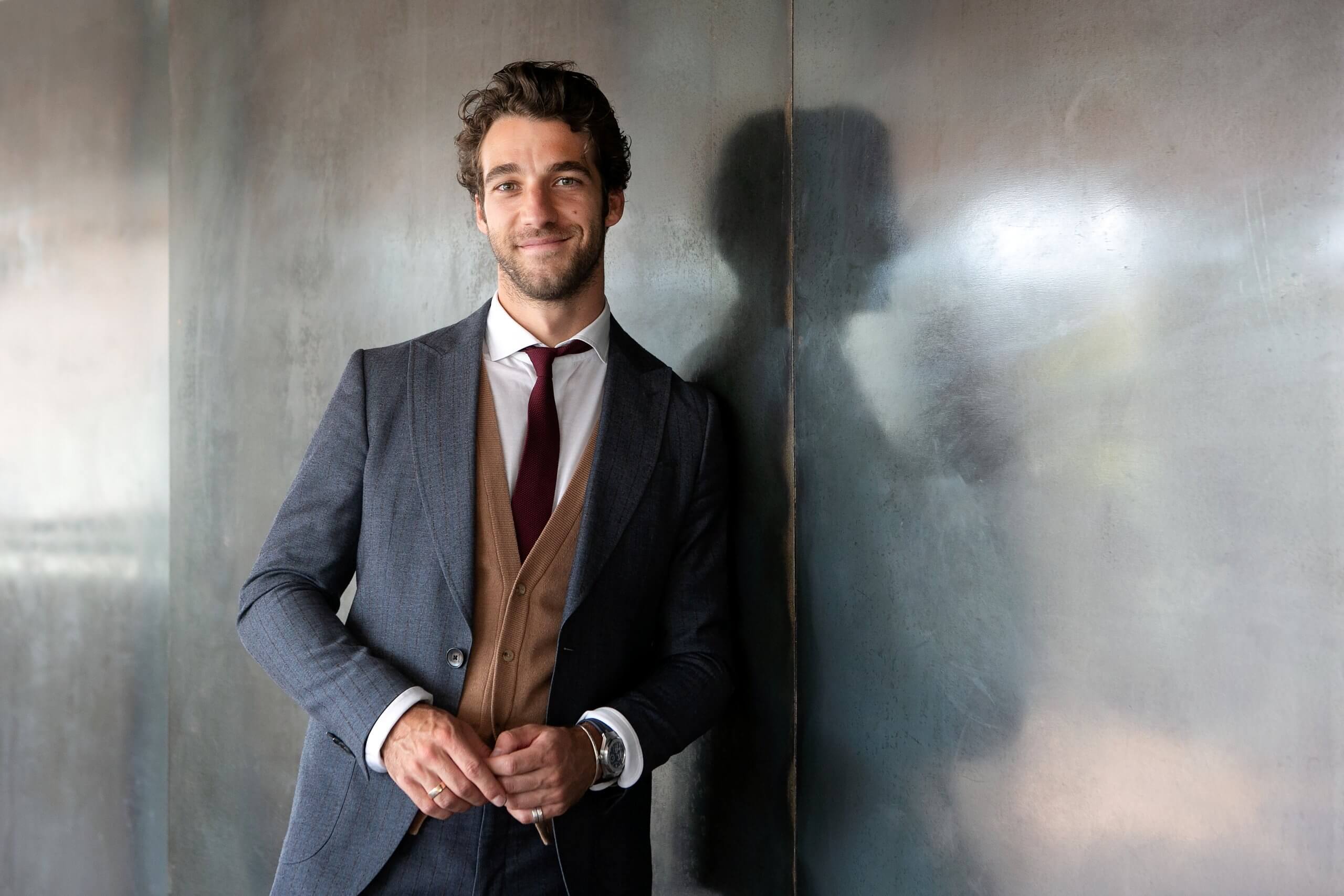 Letsmilende og varme mørkhårede Lorenzo Viotti læner op ad reflekterende væg iført gråt jakkesæt med rødligt slips.