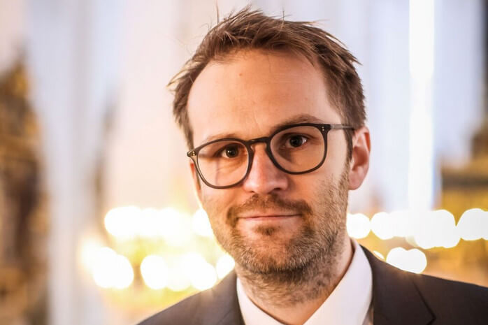 Kristian Krogsøe med sort briller og et skævt lille smil foran lys sløret baggrund.