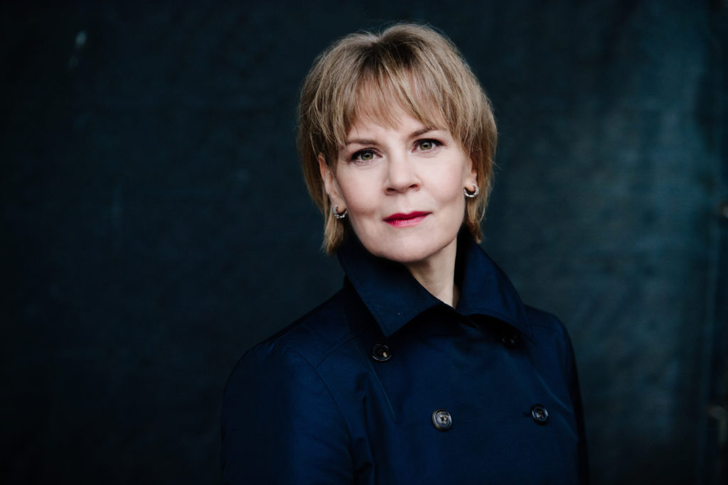 Dirigenten Susanna Mälkki med skulderkort lyst hår iført lukket mørkeblå efterårsjakke foran mørk baggrund. 