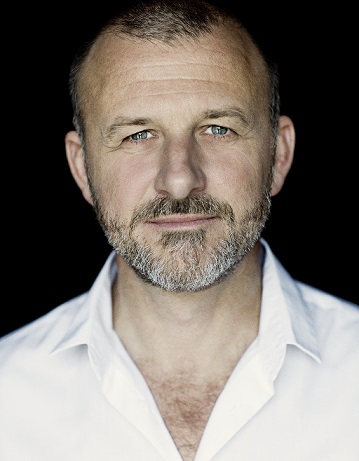 Instruktøren Nikolaj Cederholm iført hvid åbenstående skjorte med blå milde øjne og lukkede læber smiler svagt.